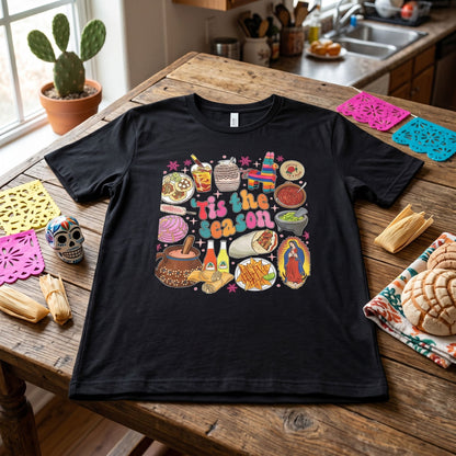 Flat lay of 'Tis the Season' t-shirt styled with seasonal accessories, highlighting print and fabric texture.